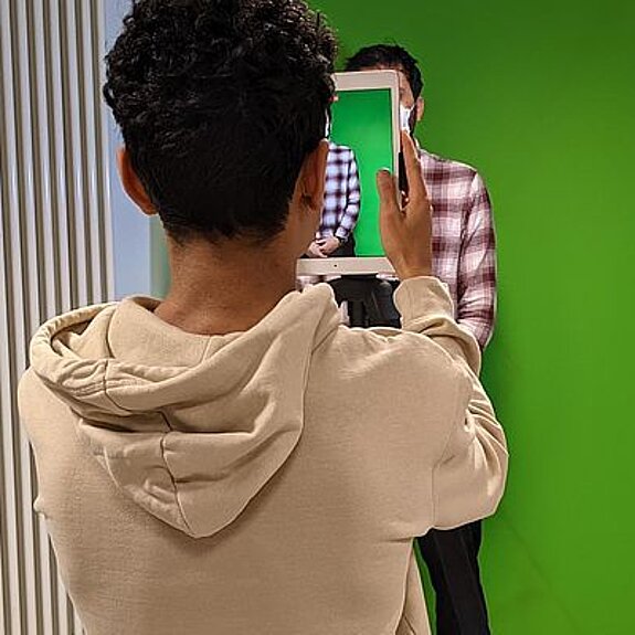 Junger Mensch fotografiert mit Tablet eine Person die vor einem Greenscreen steht.
